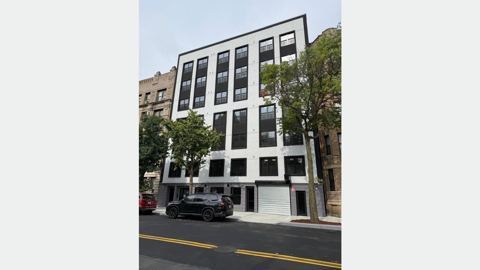 Photograph of a new seven-story apartment building with a white facade, tall black window frames in vertical bands, a gray ground floor with a metal roll-down gate and two doors, two street trees, and a black SUV and a red car parked at the curb
