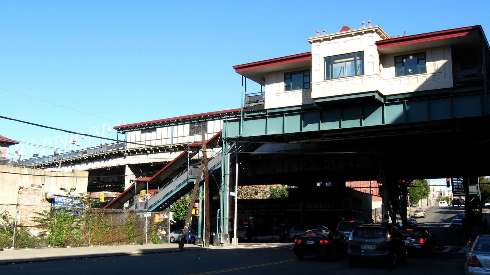A station building with a cream facade and a red roof on green steel over a wide street, a long platform canopy and street stairs at left, parked cars and traffic below, under a clear blue sky