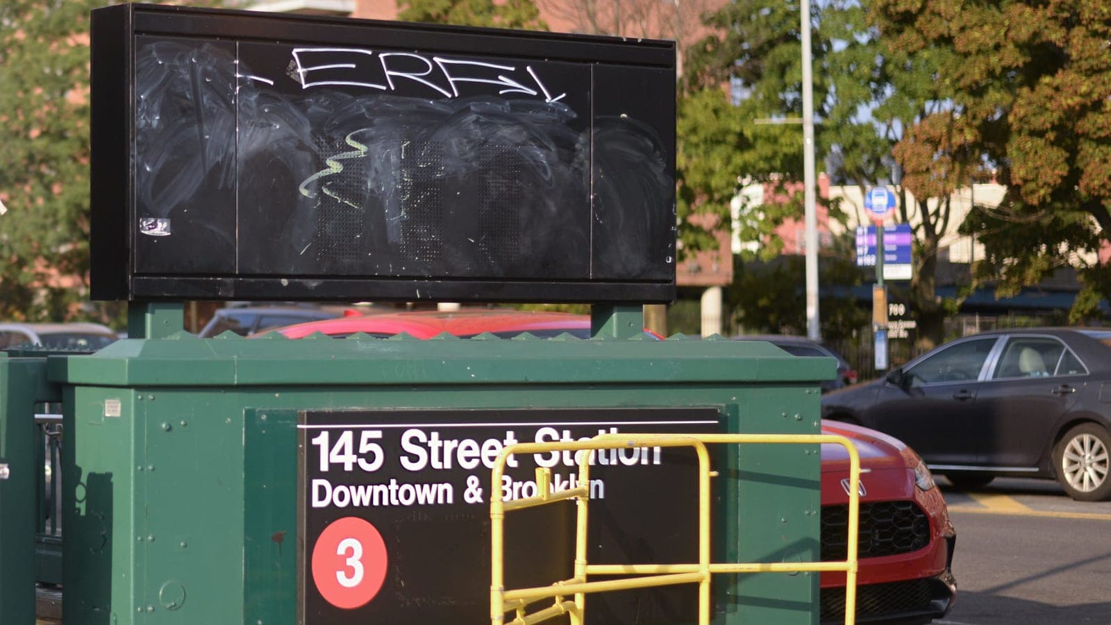Photograph of a green subway entrance kiosk with a black sign reading 145 Street Station, Downtown and Brooklyn, with a red 3 train symbol, a yellow metal barrier in front, a blank black display board with graffiti above, parked cars at right and trees and a brick apartment building behind
