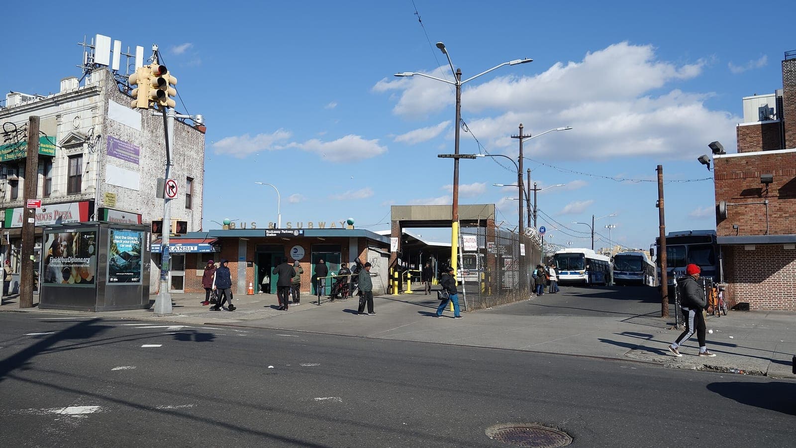 A low brick station house with a sign reading Bus and Subway and a Rockaway Parkway Station sign over two open doorways, people walking on the wide sidewalk in front, a newsstand kiosk with posters at left beside a corner building with a pizzeria, and a fenced bus loop with several city buses at right under a blue sky