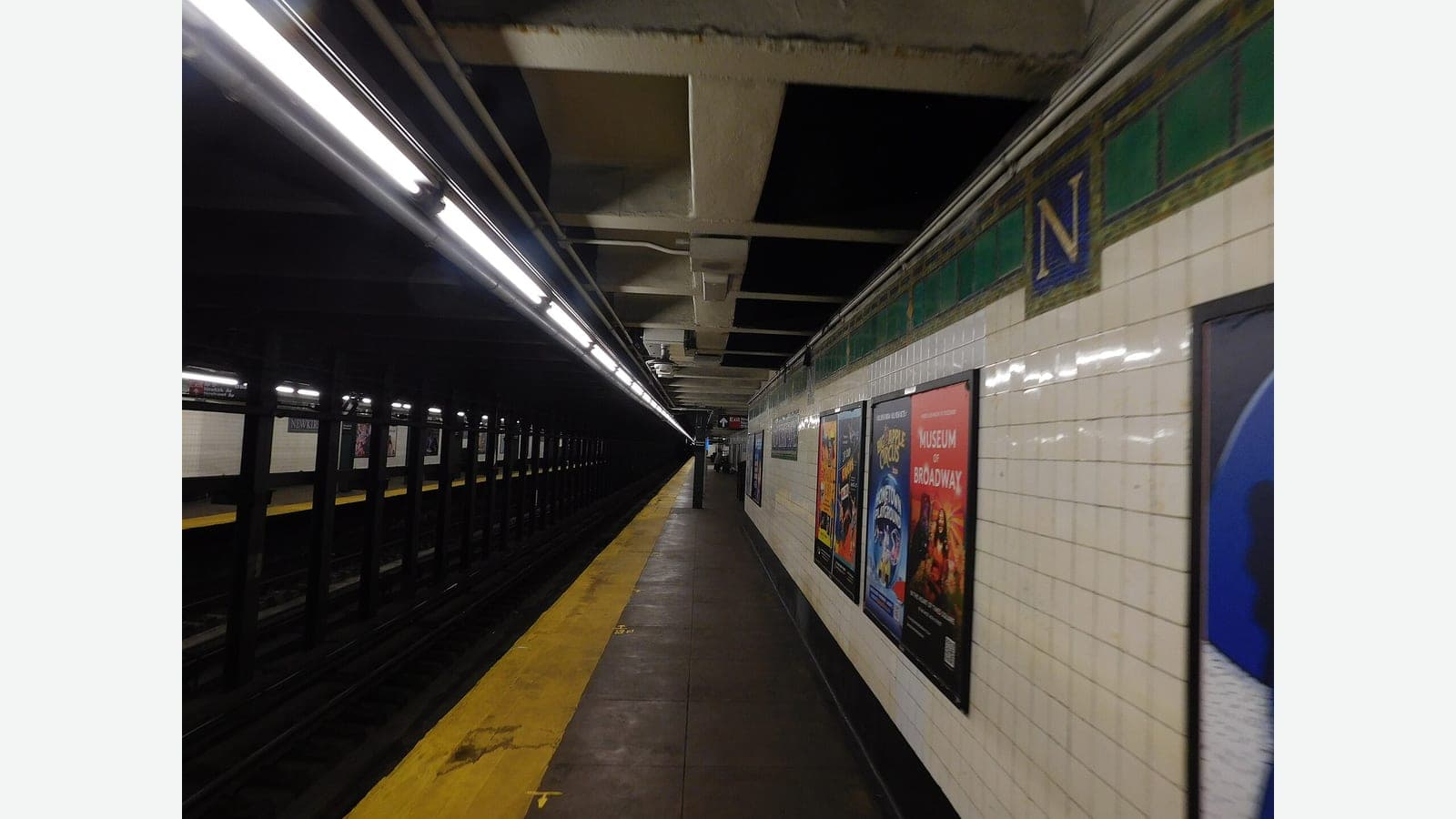 Photograph of an empty underground subway platform with a yellow safety edge, fluorescent lights on the ceiling, white tiled walls with a green tile band, a mosaic letter N plaque and posters, and the track on the left