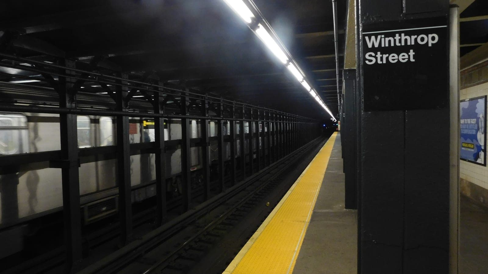Photograph of an underground subway platform with a black sign reading Winthrop Street on a dark steel column at right, a yellow tactile strip along the platform edge, a row of fluorescent lights overhead, dark steel columns between the tracks and a silver train standing on the far track at left