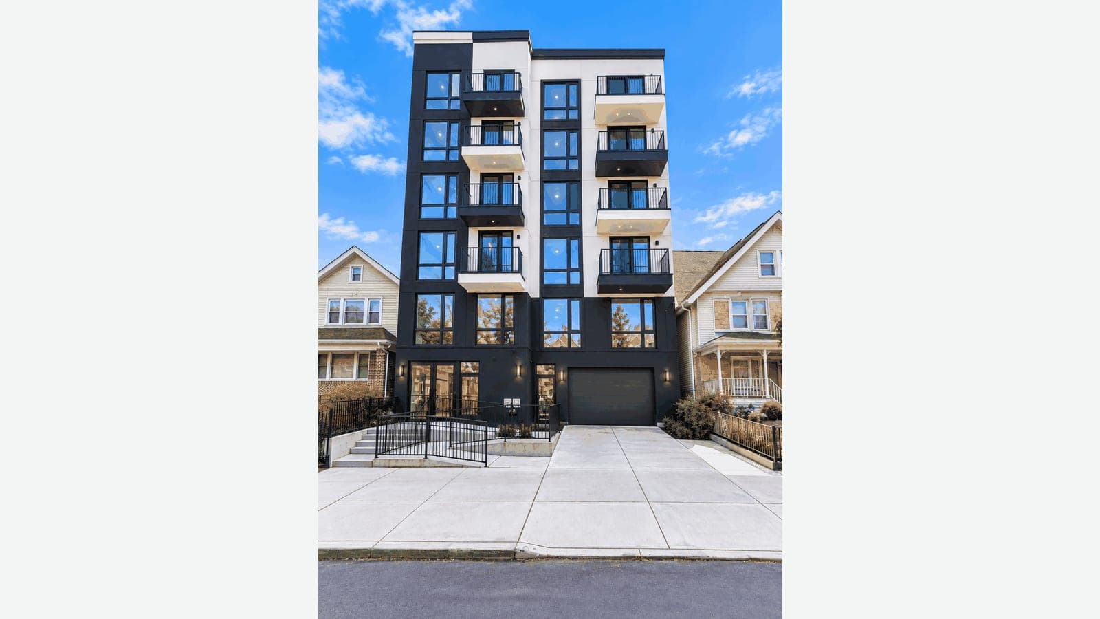 Photograph of a six-story apartment building with a black and white facade, black-framed windows and stacked balconies with black railings, between two older two-story frame houses with porches, a ramp and steps with black railings to a glass entrance on the left, a dark garage door on the right, and a wide new concrete sidewalk under a bright blue sky
