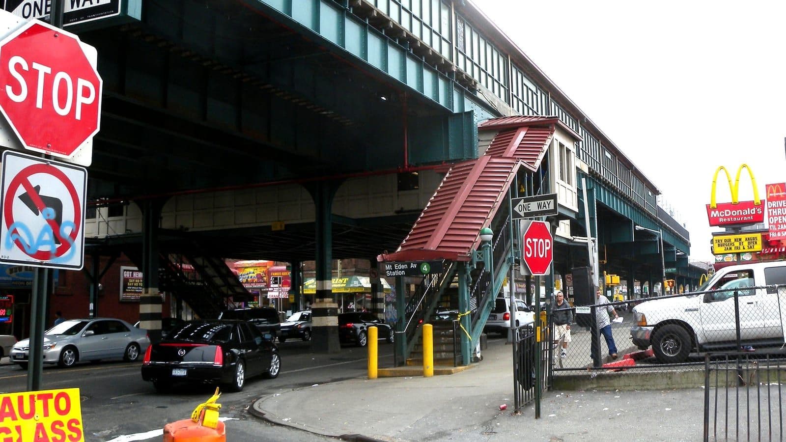 Photograph of a covered red stair climbing to an elevated green steel subway structure, with a sign reading Mt Eden Av Station and a green 4 symbol, stop signs and one-way signs, parked cars under the tracks, two men on the sidewalk and a McDonald's sign on the right