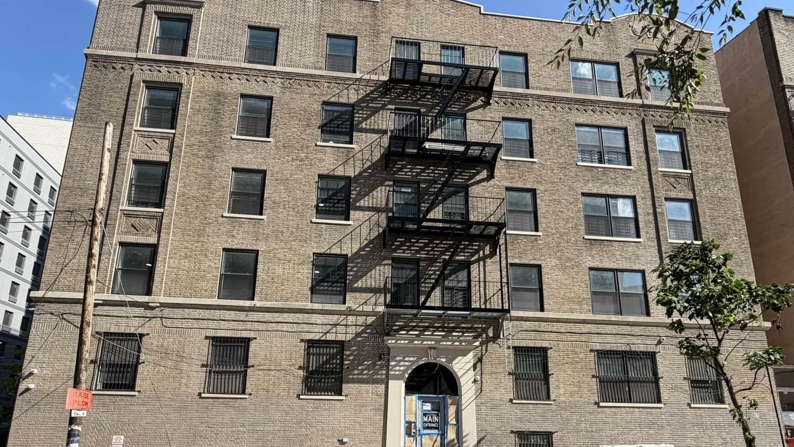 Photograph of a six-floor tan brick apartment building seen from the street on a sunny day, with a black metal fire escape down the middle of the front, new dark window frames, barred windows on the ground floor, decorative brick patterns under the roofline, an arched main entrance with a temporary blue-framed door marked MAIN ENTRANCE, a wooden utility pole at left and a young street tree at right