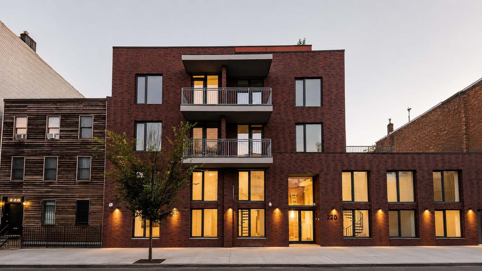Dusk photograph of a dark red brick apartment building: a four-story section at left with two stacked balconies in the middle, a lower wing at right with a railing along its roof, tall lit ground-floor windows with spiral stairs visible inside, a glass entrance with a house number beside it, a young street tree on the sidewalk, and a wood-sided three-story house at left