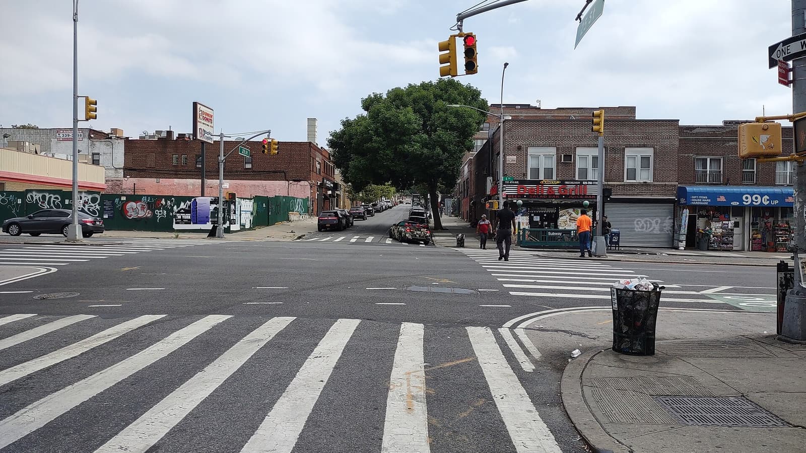 Wide crosswalk at a four-way intersection under a cloudy sky, with traffic lights, a green 25 St street sign, a corner deli and a 99 cent store in two-story brick buildings, a green subway entrance marked Subway, and a fenced lot covered in graffiti across the street