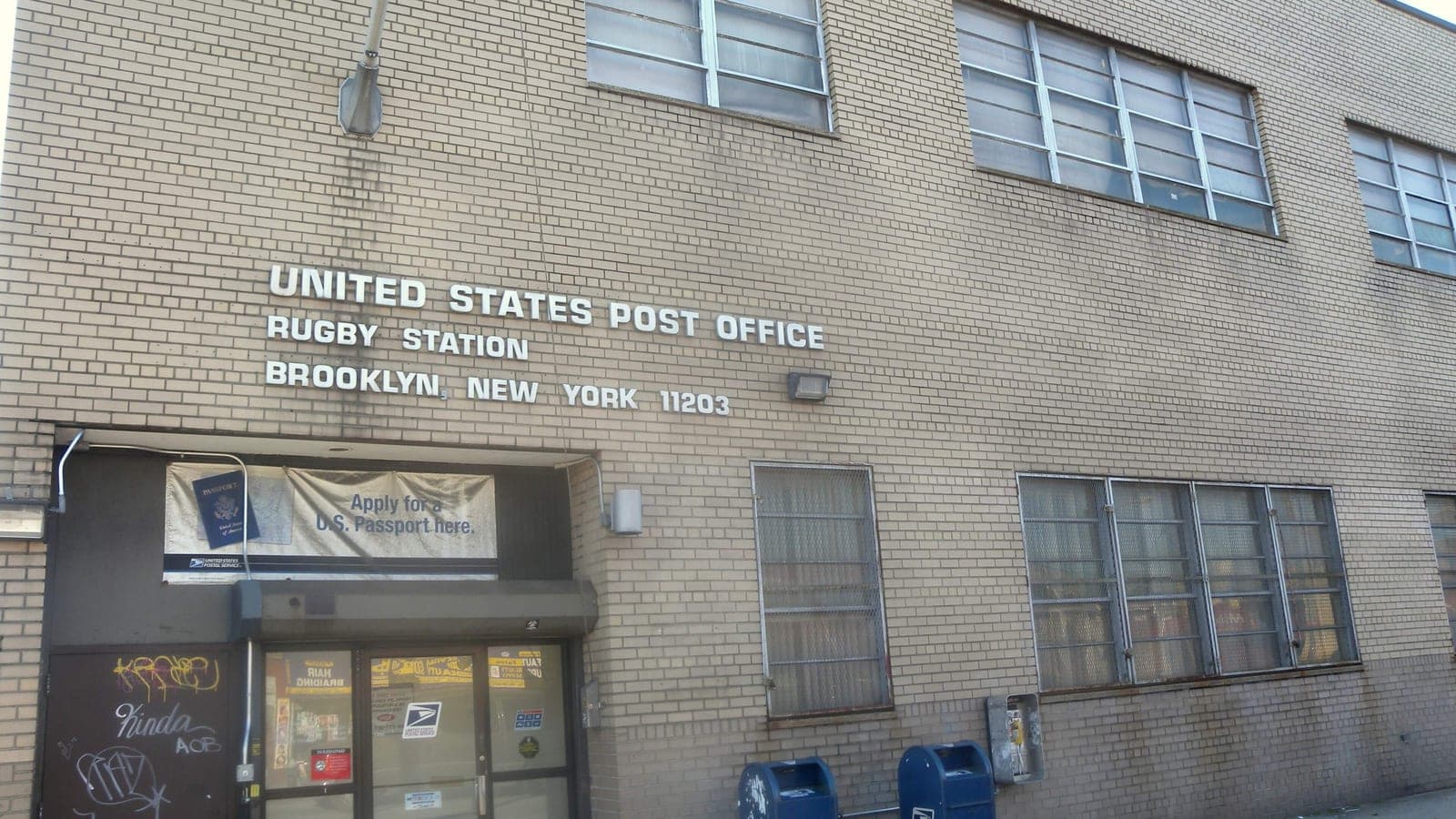 Photograph of a tan brick two-story post office with white letters reading United States Post Office, Rugby Station, Brooklyn, New York 11203, a banner reading Apply for a U.S. Passport here over the glass entrance doors, graffiti on a side door, and two blue mailboxes on the sidewalk