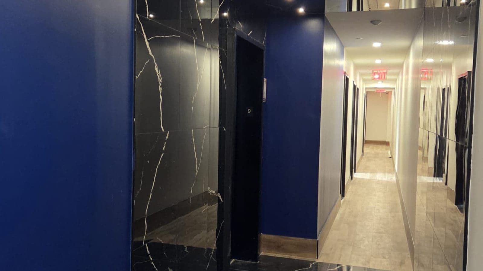 A building hallway with a dark blue wall at left, black marble-look panels around a black elevator door marked 9, and at right a mirrored wall beside a long corridor with wood-look floor tiles, recessed ceiling lights and red exit signs