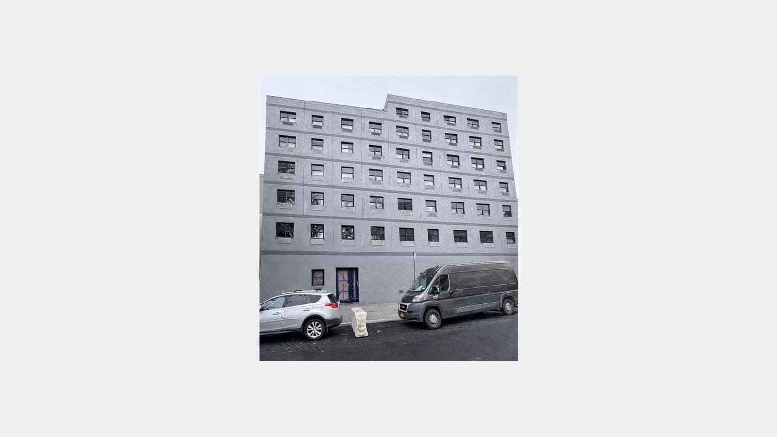 Photograph of a plain gray seven-story apartment building with rows of square black windows and wall vents under each window, a front door covered with protective board, and a silver SUV and a dark van parked at the curb in front