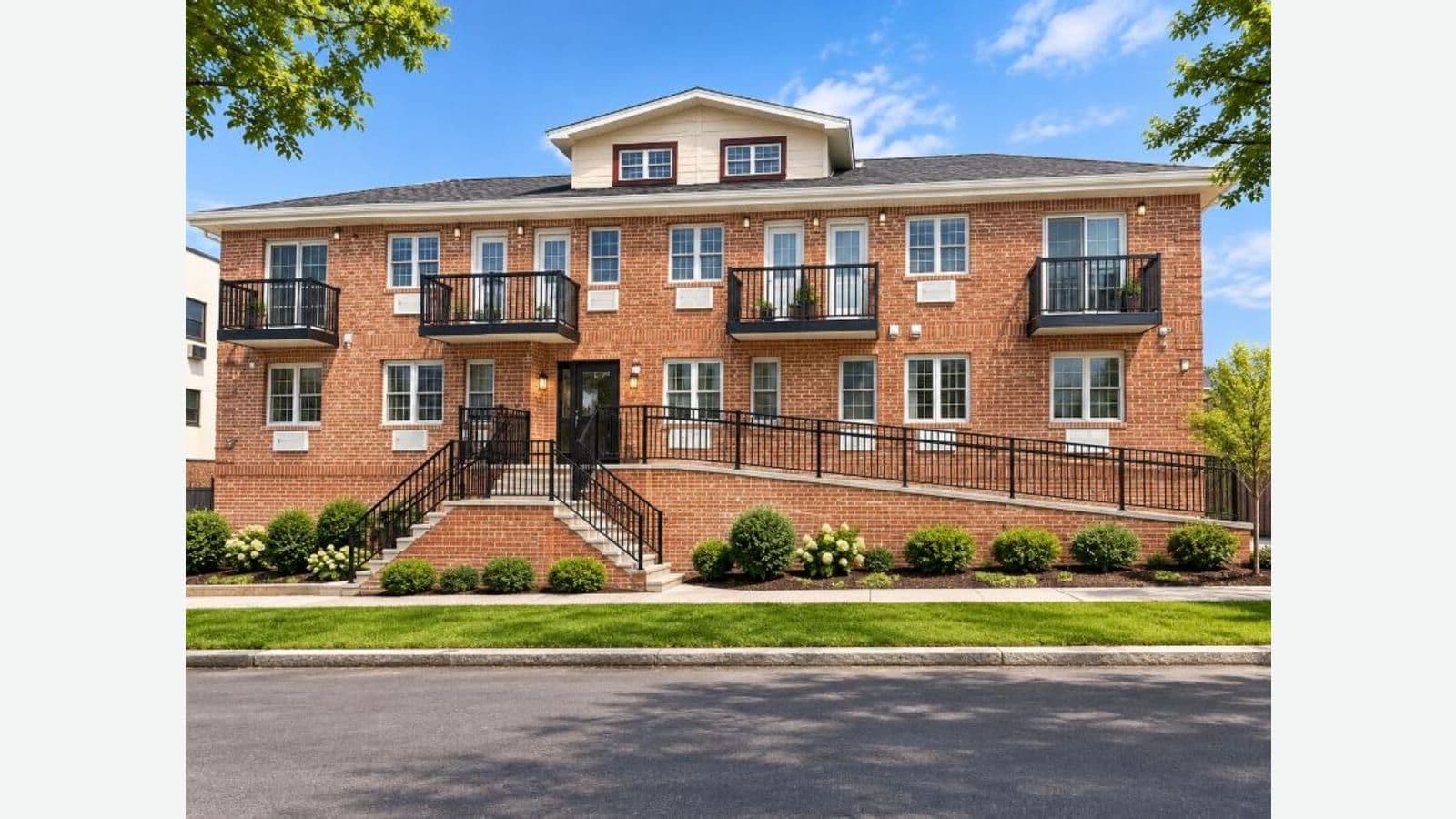 A wide red brick apartment building under a blue sky, with two rows of white-framed windows below a grey roof and a cream-colored dormer holding two red-framed windows; four small balconies with black railings on the upper row, a black front door at the top of brick stairs, and a long ramp with black railings running along the front to the entrance, with clipped shrubs and white hydrangeas in a bed along the sidewalk, a strip of lawn and an empty street in front