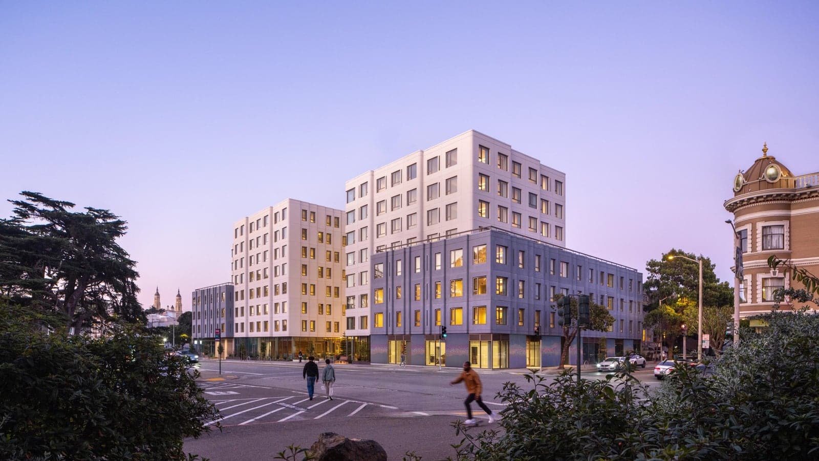 An eight-story white apartment building with a lower gray wing at a street corner at dusk, lit from inside, with people crossing the street, trees in the foreground and a Victorian building at right