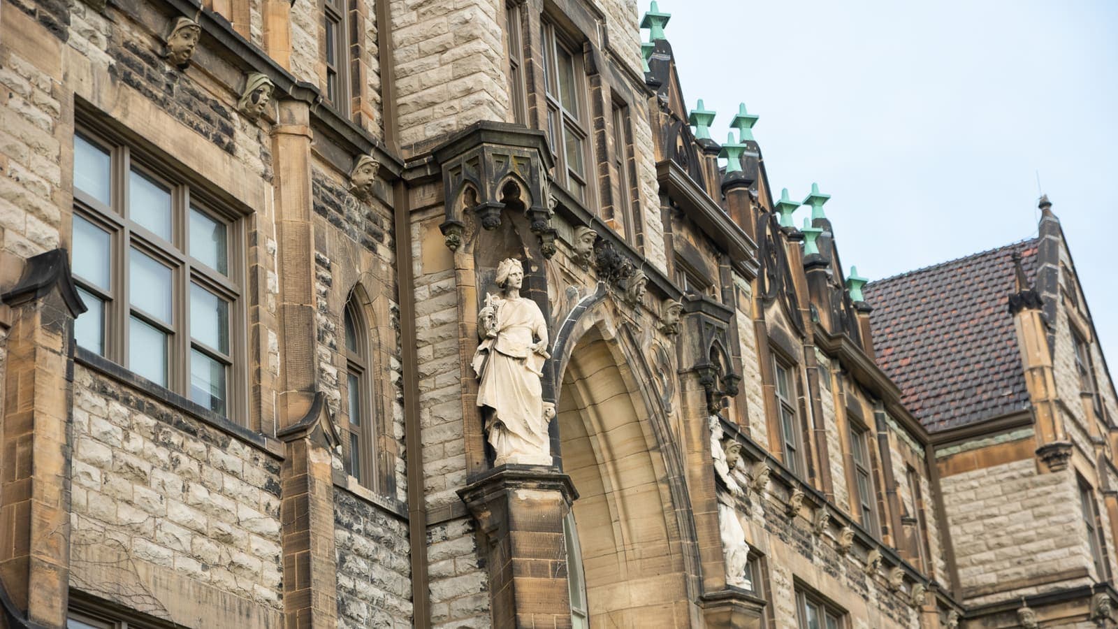 Stone facade of a Gothic Revival school building with a statue in a carved niche beside a pointed arch, copper finials along the roofline and a tiled gable