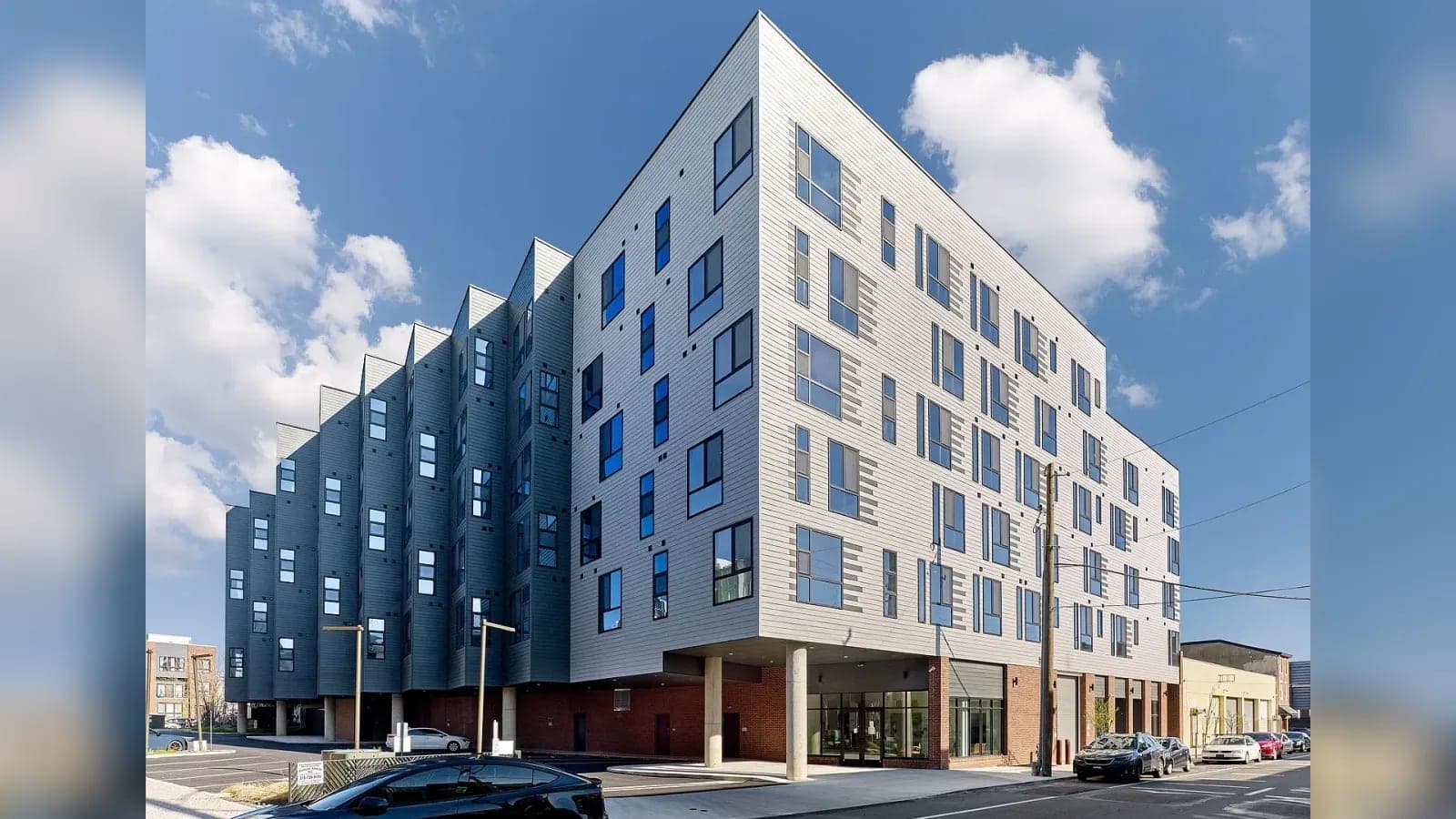 Six-story apartment building with pale and dark gray horizontal siding, a sawtooth side wall, a red brick ground floor on columns and cars parked along the street under a blue sky