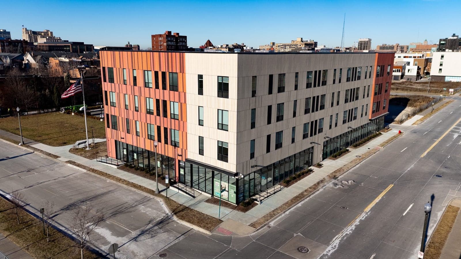 Aerial photograph of a four-story apartment building with cream panels, orange and red ends and a glass ground floor at a street corner, with a flag on a pole beside it
