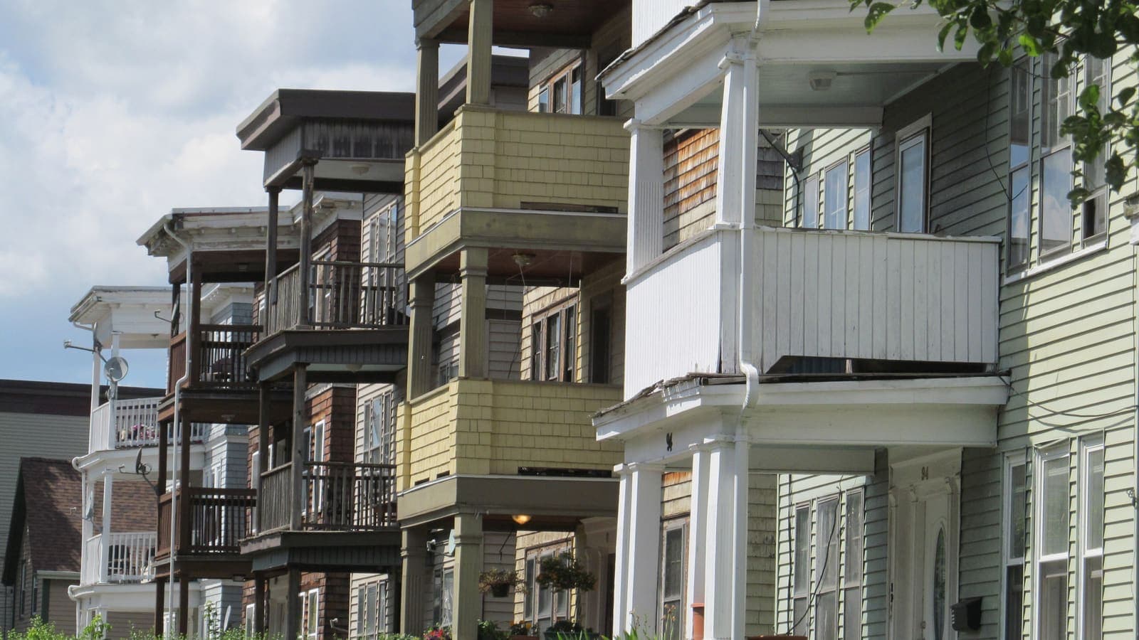 A row of wooden three-storey houses with stacked front porches, in yellow, brown, white and pale green, seen at an angle along a street