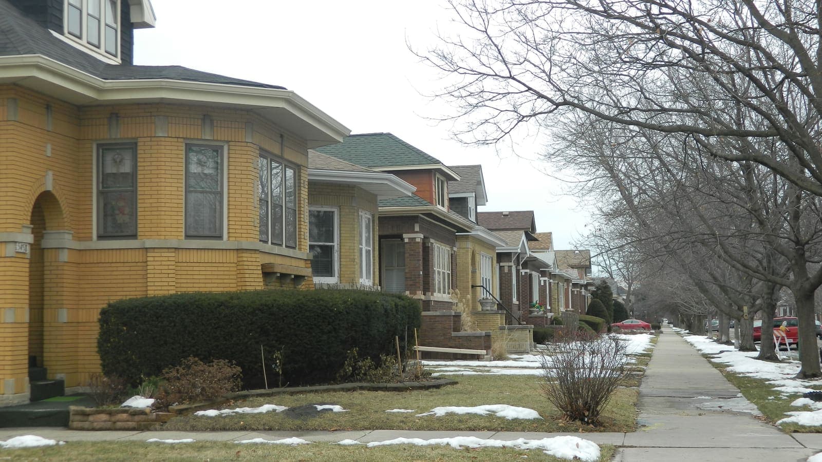 A row of single-storey yellow and brown brick bungalows with small front lawns along a tree-lined sidewalk, with patches of snow on the grass