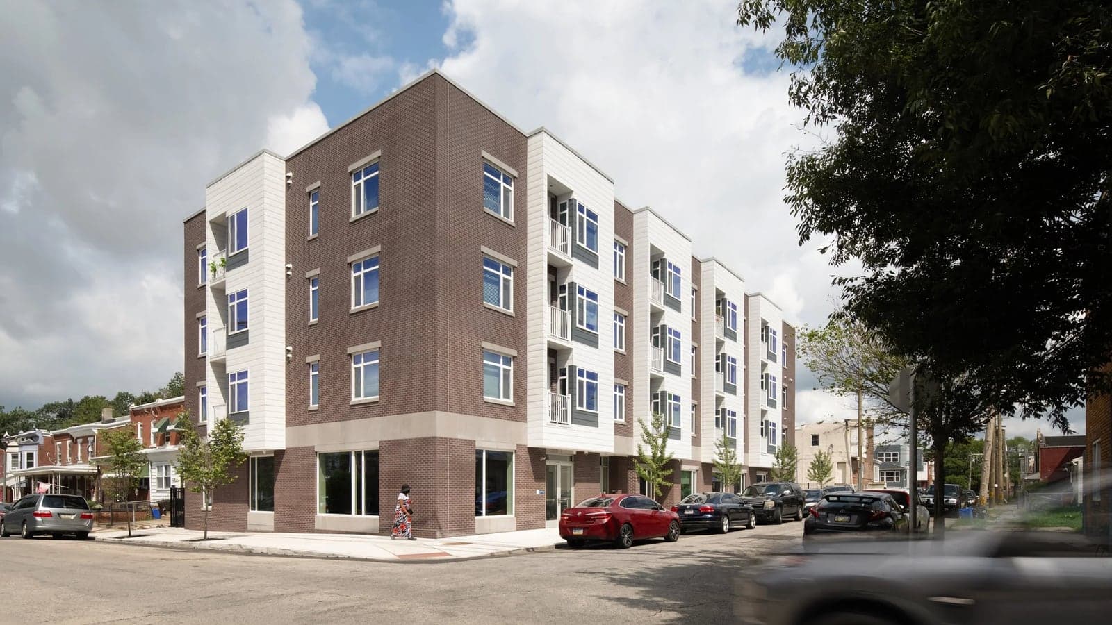 Four-story corner apartment building in brown brick and white panels with balconies, large ground-floor windows and cars parked along the street under a cloudy sky