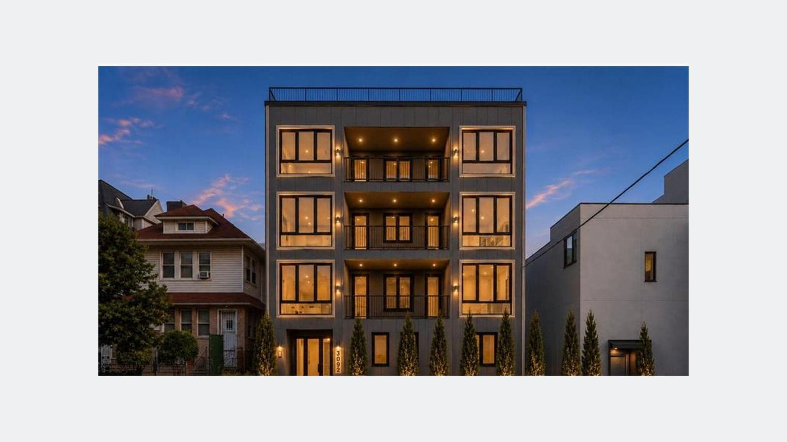 A four-story apartment building with a flat roof and rooftop railing, faced in dark grey panels, seen from the street at dusk. Its large windows and three stacked balconies in the middle are lit warm yellow. A row of narrow evergreen shrubs lit from below lines the front, the number 3092 is beside the glass entrance door, a two-story frame house stands at the left and a plain grey building at the right.