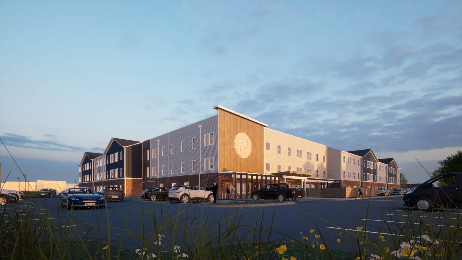 Rendering at dusk of a three-story apartment building with gray and tan siding, a wood-clad entrance bay with a round emblem and cars parked in front