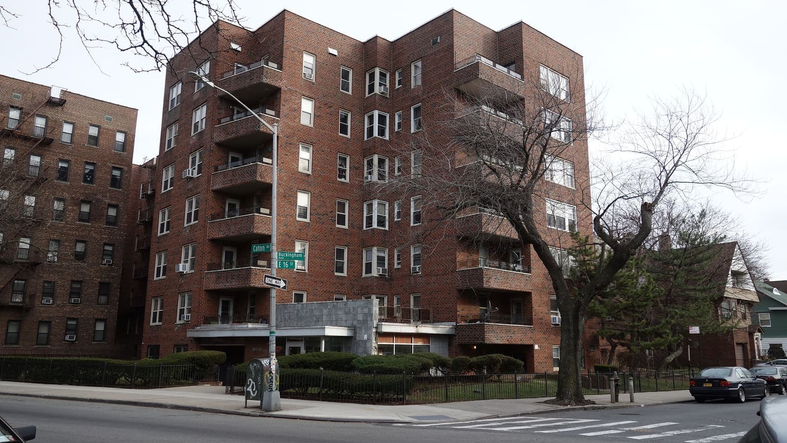 A seven-story red-brick apartment building with corner balconies behind a low iron fence, with street signs for Caton Avenue, Buckingham Road and East 16th Street on a lamp post in front.