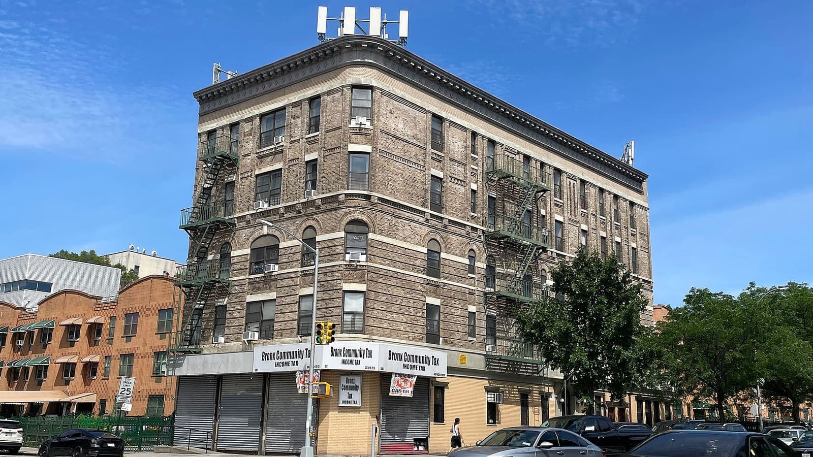 A five-story brick corner apartment building with green fire escapes and shuttered ground-floor shops, under a blue sky, with parked cars in front.