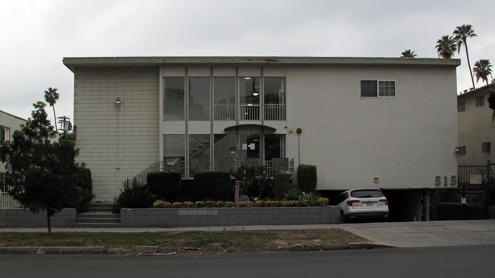 A two-storey stucco apartment building with a glass-fronted stair hall, a planted front wall and a car parked under the upper floor