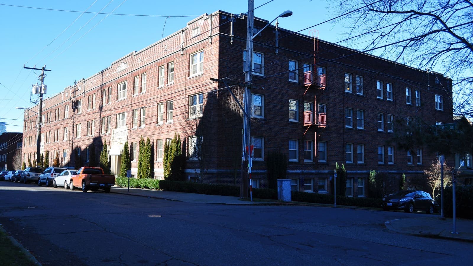 A long three-storey red-brick apartment building on a street corner, with parked cars along one side and bare tree branches at the right
