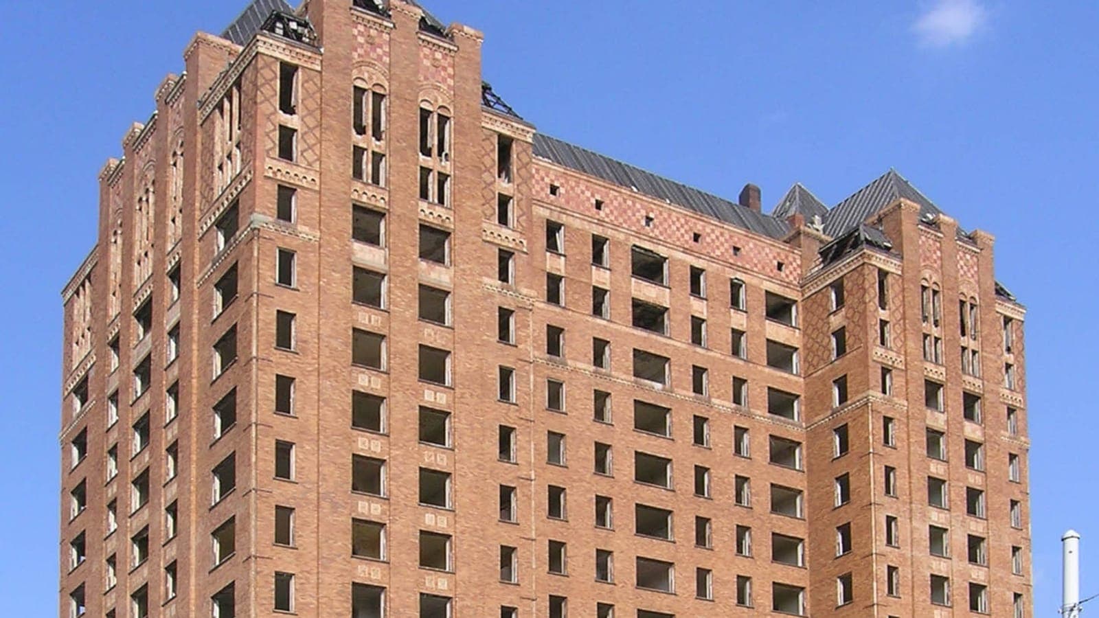 Photograph of the upper floors of an orange brick tower with rows of empty window openings and steep roofs at its corners, against a blue sky