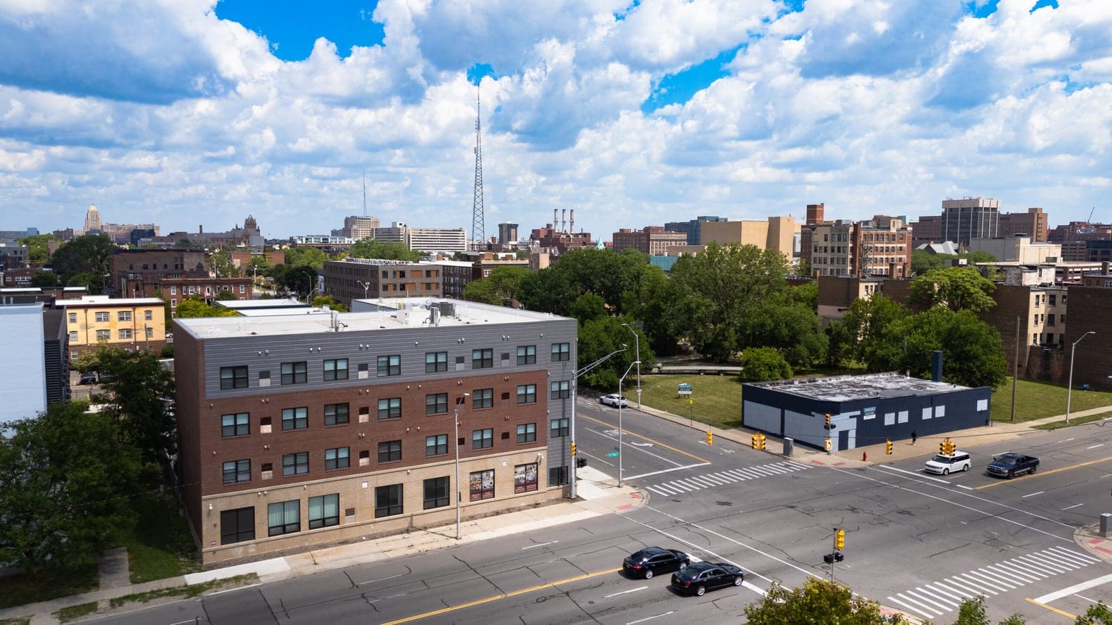 Aerial photograph of a four-story apartment building with dark red brick, a gray top floor and a pale ground floor, at a street corner with traffic signals and trees behind it