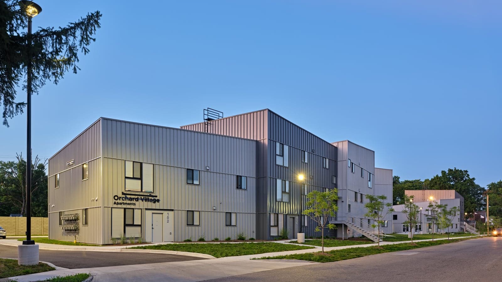 Photograph at dusk of a gray metal-clad apartment building of two and three stories lettered Orchard Village Apartments, with lit lamps, young trees and more buildings behind it