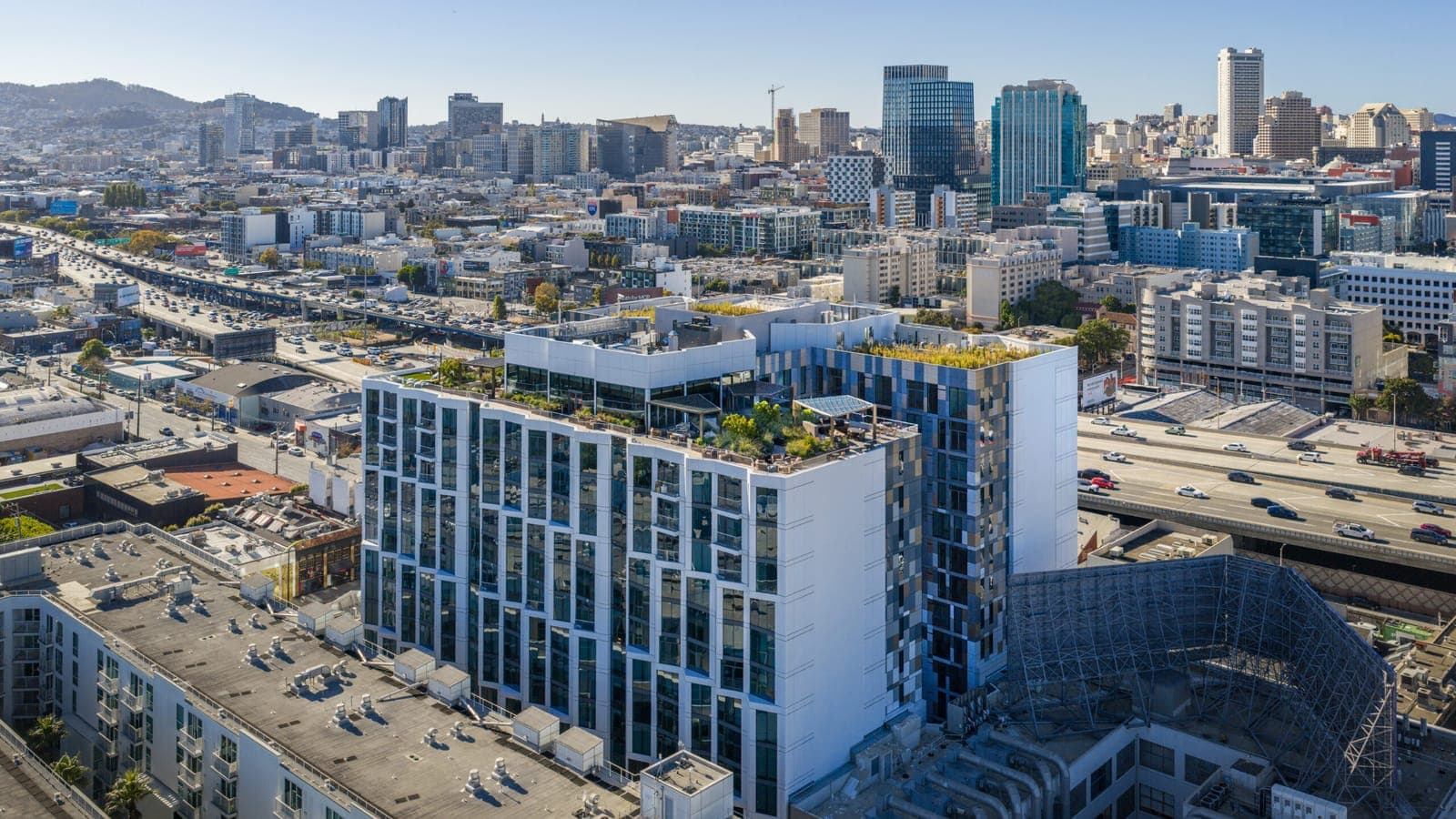 Aerial photograph of a 16-story white and glass apartment building with planted roof terraces, an elevated freeway running past it and the towers and hills of San Francisco behind