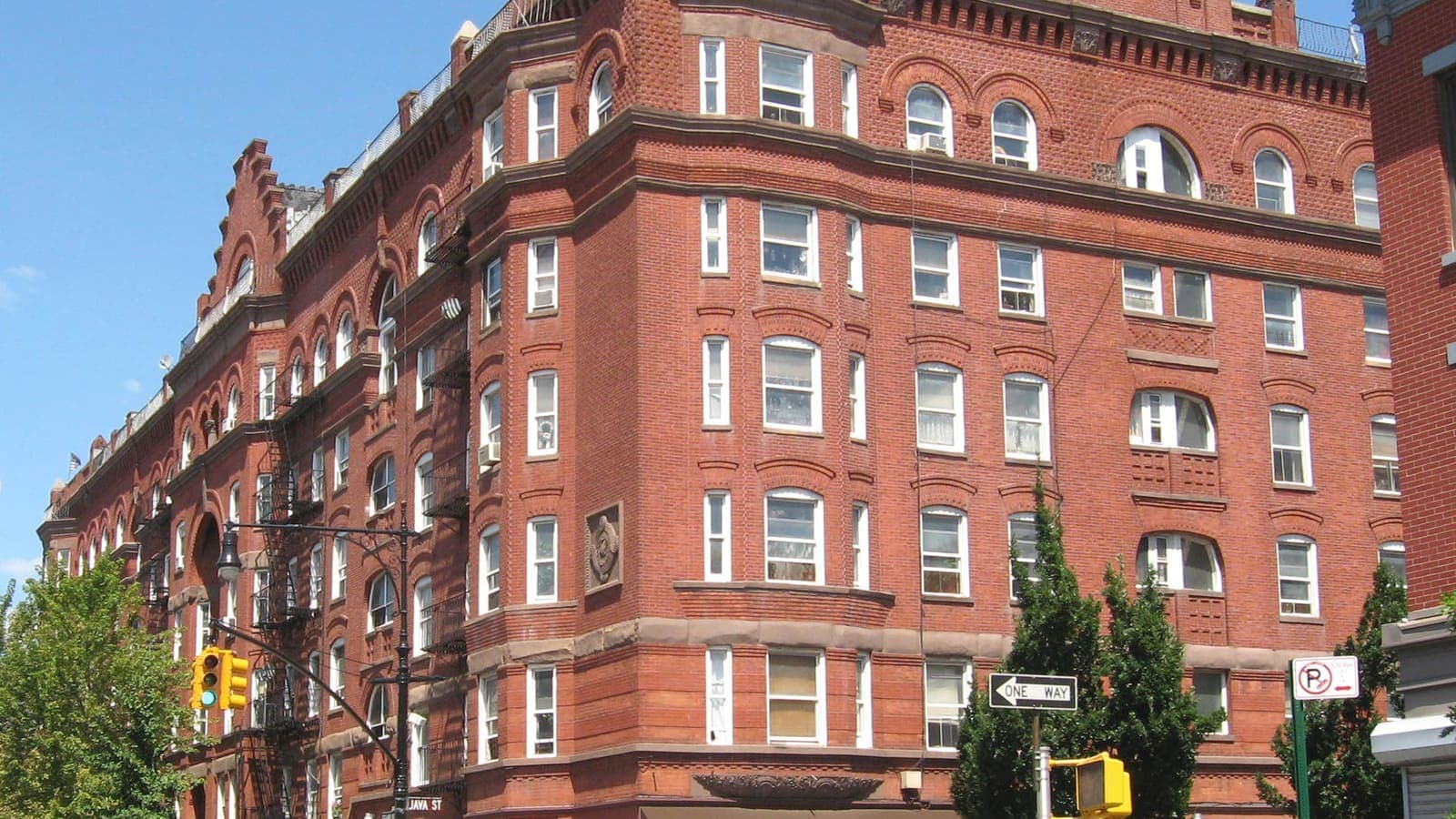 A six-story red brick apartment building with arched windows, bay fronts and fire escapes at a street corner under a blue sky