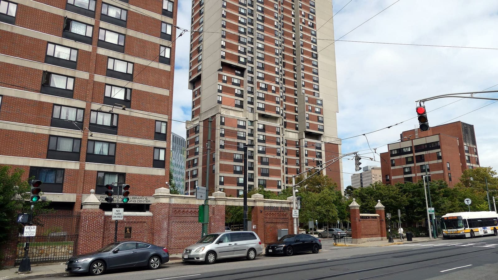Red brick apartment towers and a lower brick building behind a brick gateway marked Mission Park, with parked cars and a city bus on the street in front