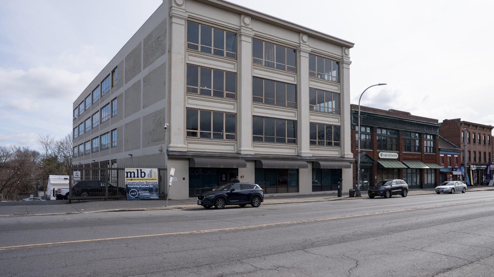 A four-story cream-colored concrete building with tall grouped windows and classical pilasters on a city avenue, with a builder's banner on the fence of the parking lot beside it