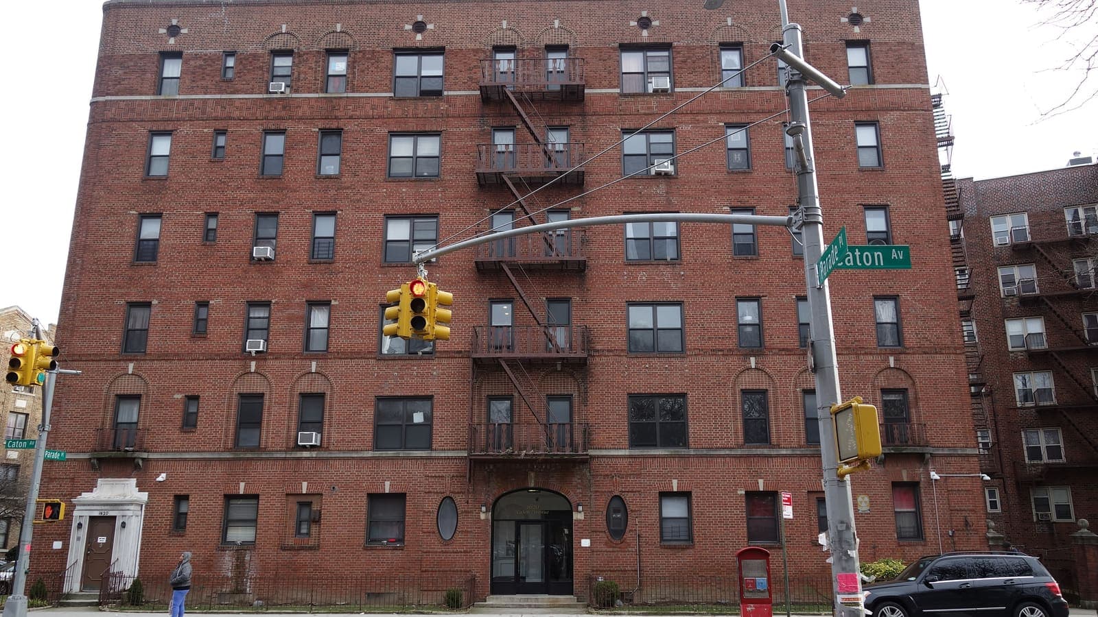 A six-storey red-brick apartment building with fire escapes on a street corner, street signs reading Caton Av and Parade Pl, a traffic light in front and a person walking past