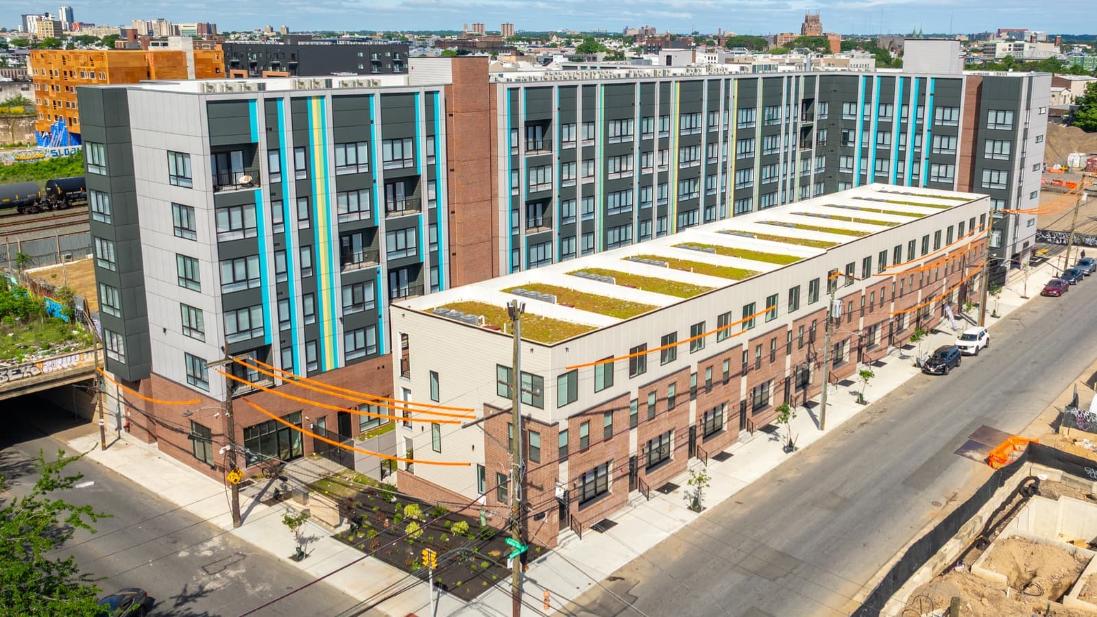 Aerial photograph of a six-story apartment building with dark gray and pale panels and blue and yellow vertical stripes, behind a row of three-story brick and white buildings with planted roofs along a street, with rail tank cars on tracks at the left
