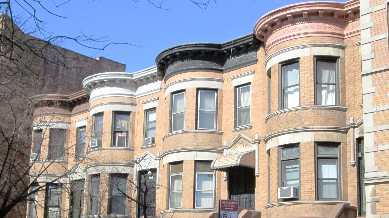 A row of two-story tan brick houses with rounded bay windows, carved cornices and window air conditioners on a city street