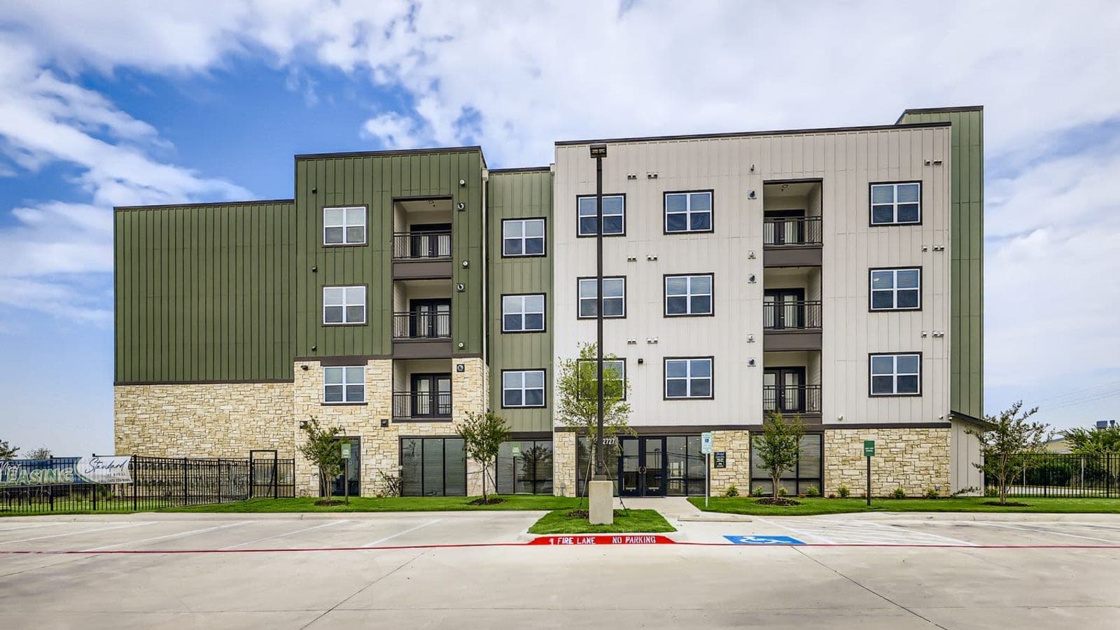 Four-story apartment building with green and cream metal siding over a stone base, balconies and a glass entrance, seen across a parking lot with a fire lane