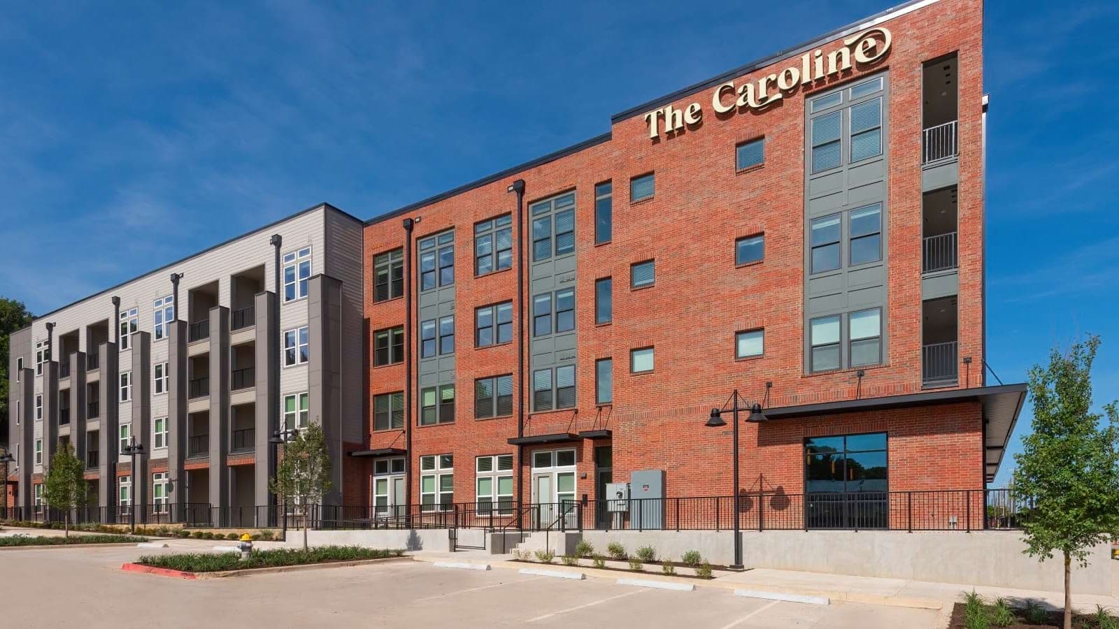 Four-story apartment building in red brick and grey panels with a sign reading The Caroline on the corner, seen across an empty parking area