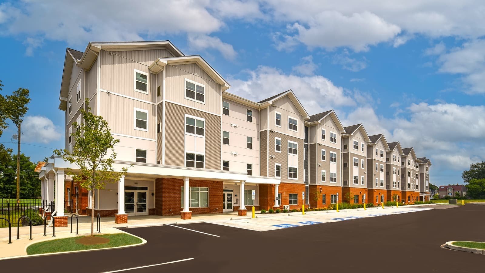 A four-story apartment building with beige and gray siding, a brick ground floor, gabled roof sections and a covered entrance with white columns, seen across its parking lot