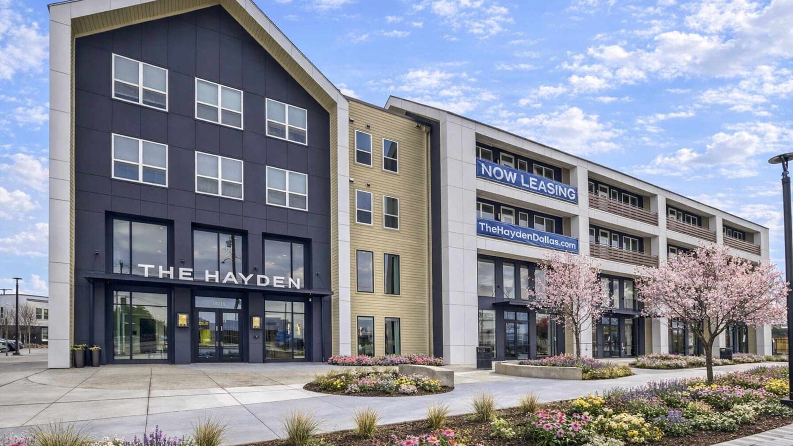 Four-story apartment building with dark and tan siding, a sign reading The Hayden over glass doors and banners reading Now Leasing, behind a planted walkway