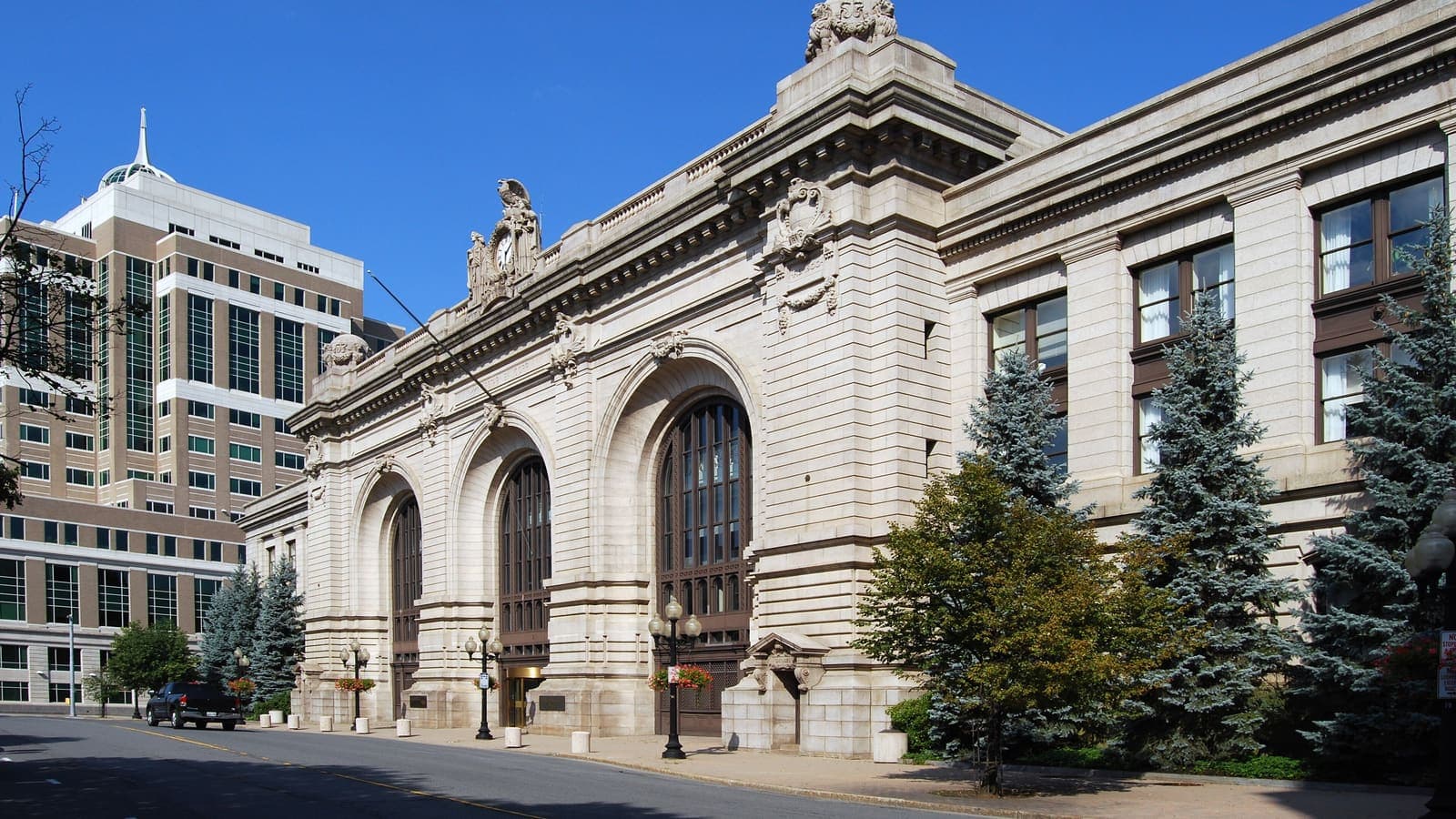 A long three-story stone building with three tall arched windows, carved figures on the roofline and evergreens along the sidewalk, beside a glass office tower