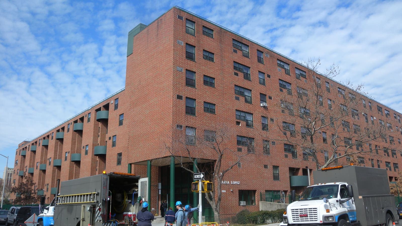 A six-story red brick apartment building with green balconies on a street corner, with two utility trucks and workers in blue hard hats on the street in front