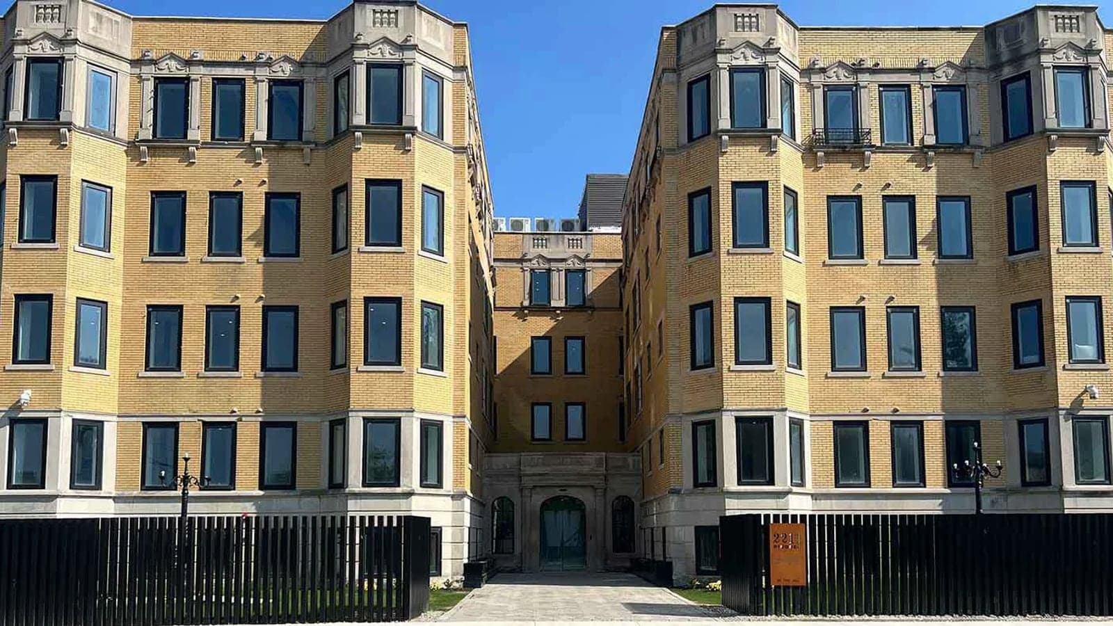 Photograph of a four-story yellow brick apartment building with bay windows and carved stone trim, its two wings facing each other across an entry court behind a black fence