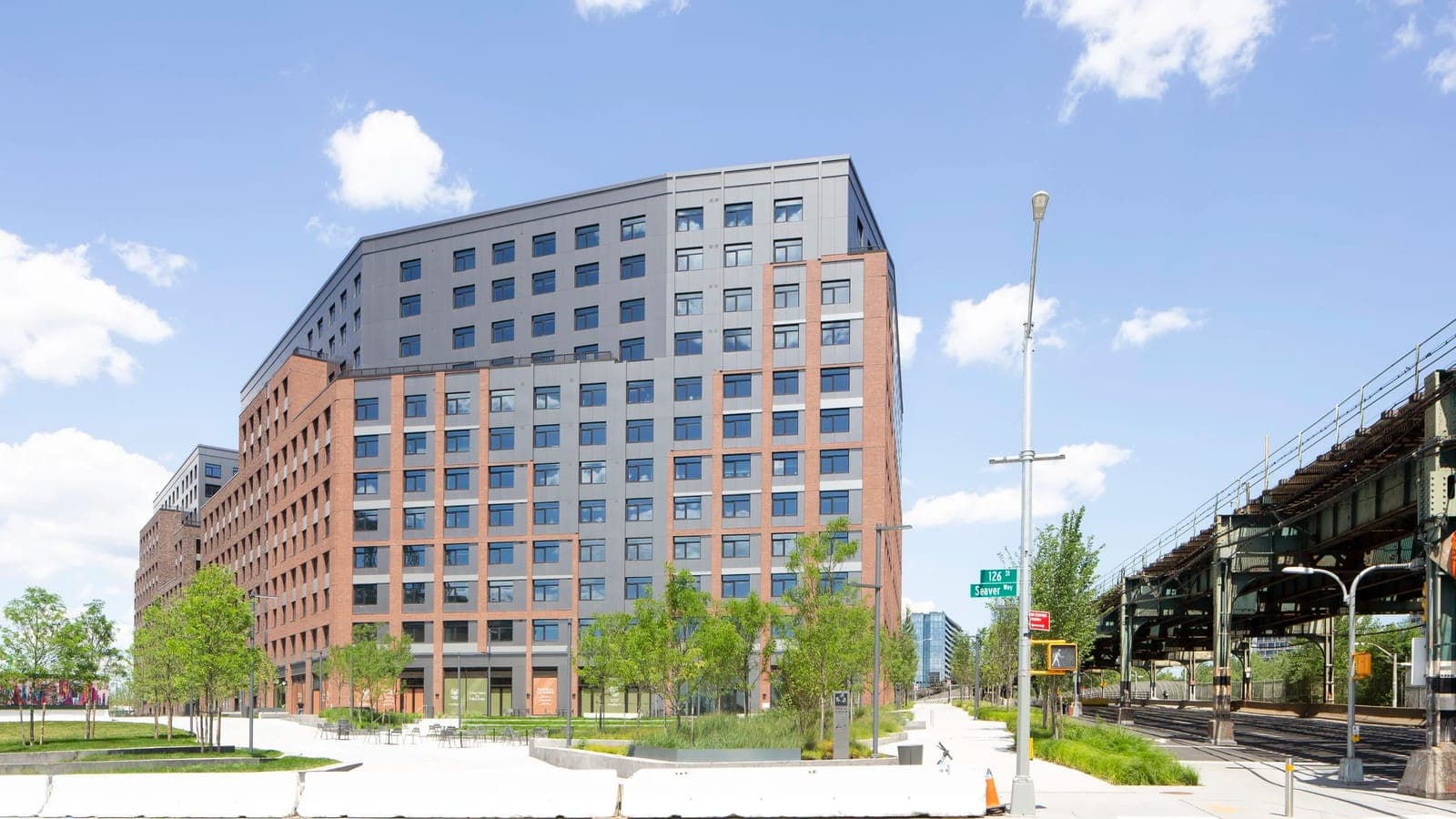 Photograph of a tall apartment building with red brick lower floors and dark grey panels above, behind a plaza with young trees, planted beds and cafe tables, with white concrete barriers in the foreground, a street sign reading 126 St and Seaver Way, and the steel viaduct of a subway line at the right under a blue sky