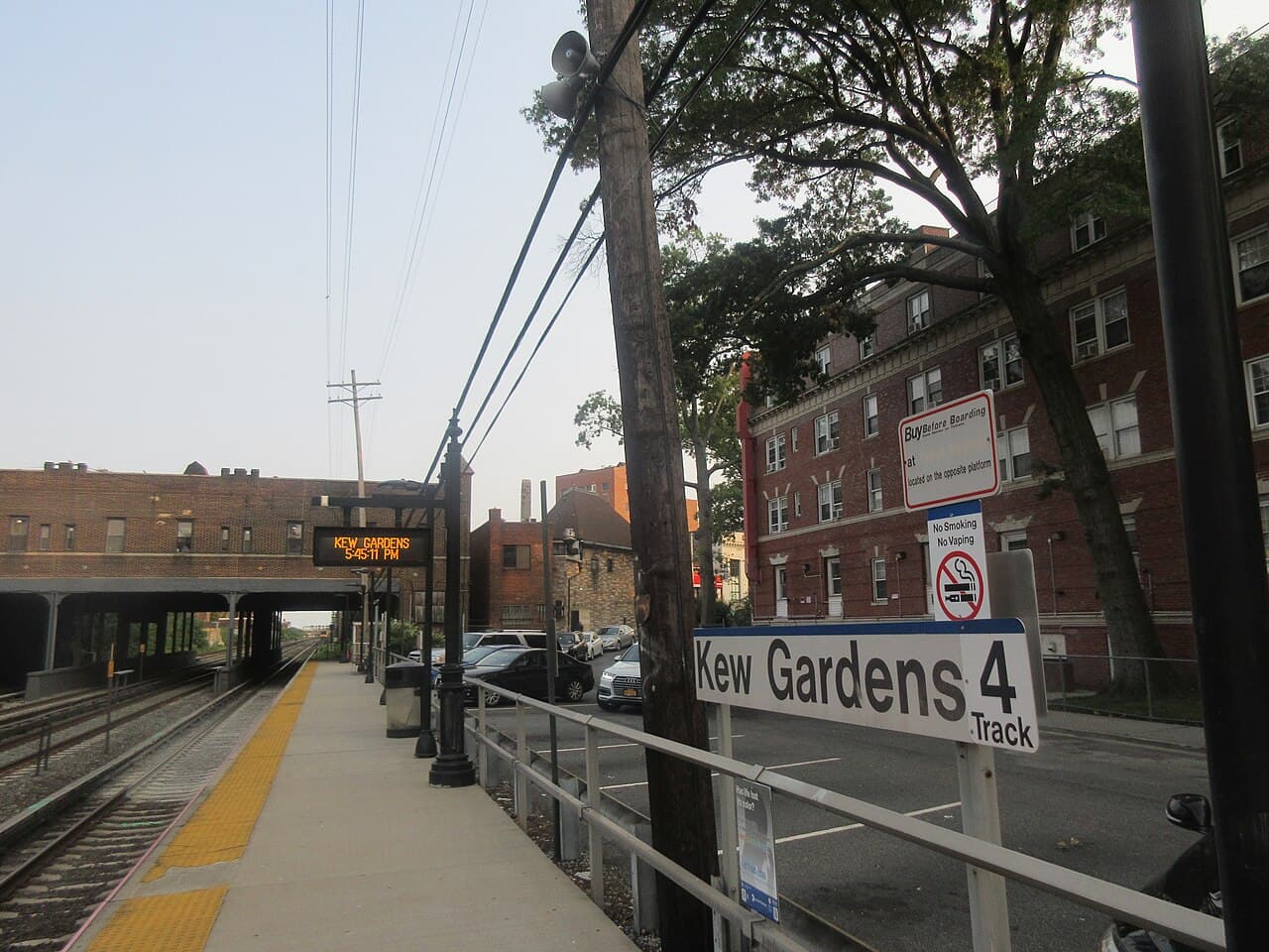 Kew Gardens LIRR eastbound platform, with the Lefferts Boulevard bridge ahead and an apartment building beside the tracks.
