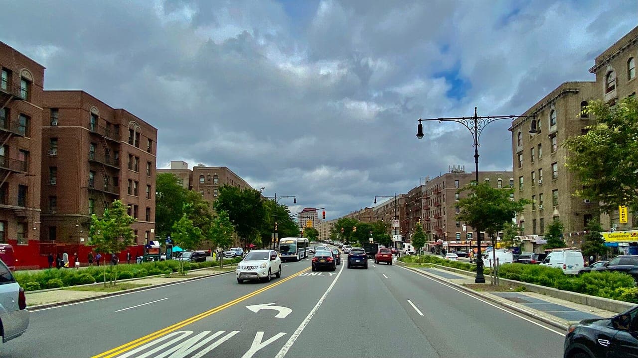Brick apartment buildings line both sides of the Grand Concourse in the Bronx, with cars and a city bus on the tree-lined boulevard under heavy clouds