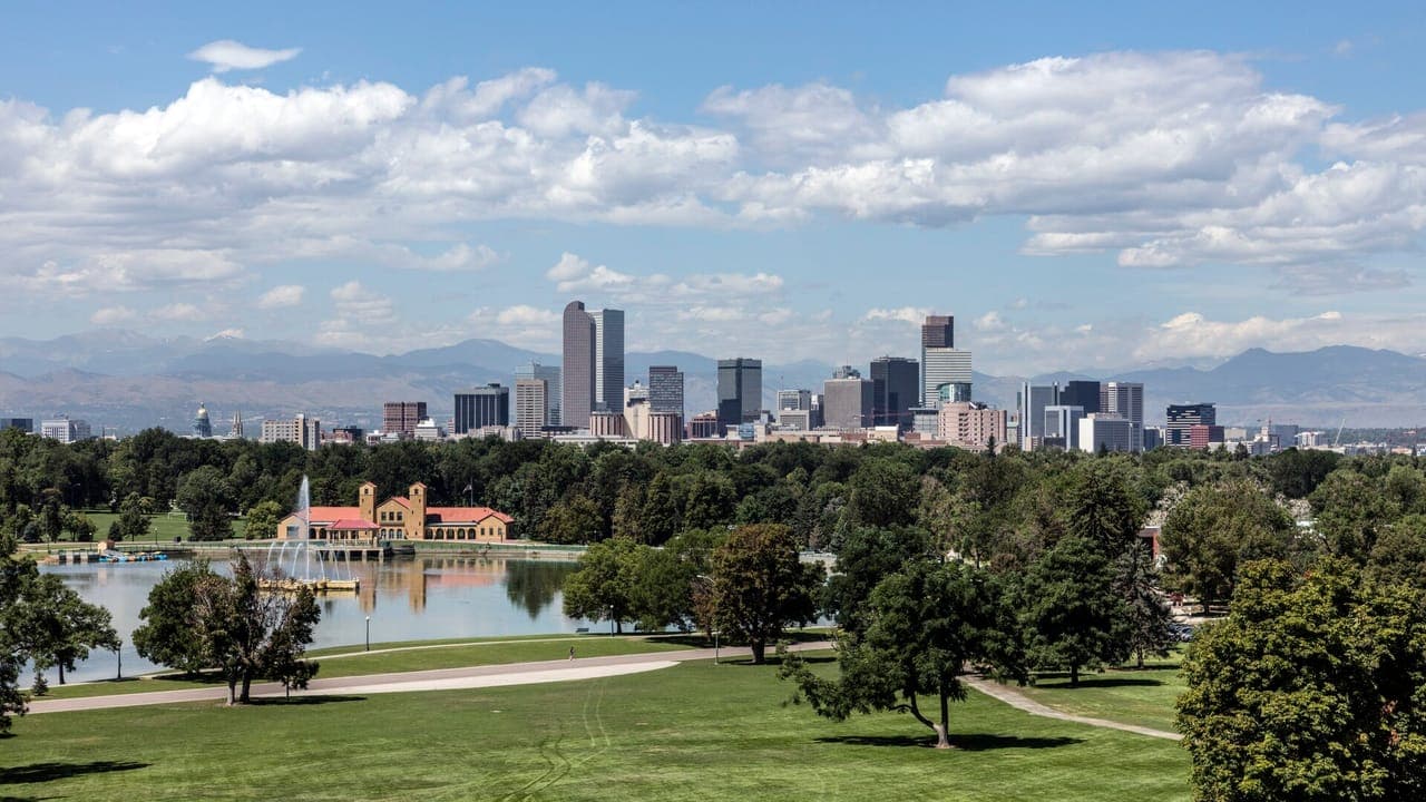 The downtown Denver skyline against the Front Range, seen across the lawns, trees and boathouse lake of City Park under a blue sky with white clouds