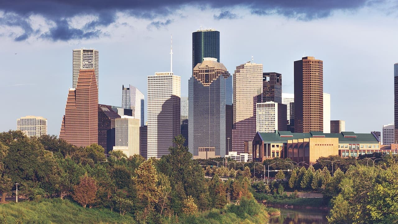The downtown Houston skyline in late-afternoon sun, seen from the west over the trees and water of Buffalo Bayou